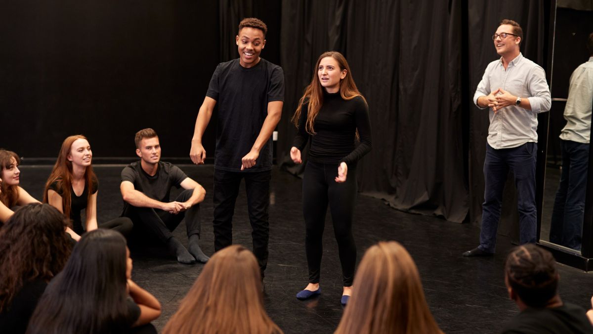 A drama class sitting on the floor of a black box studio watching two students work a scene, the teacher standing to one side