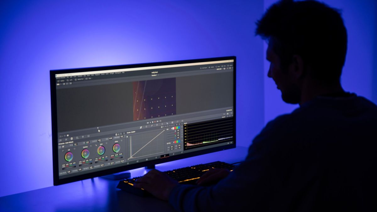 An editor in silhouette against a wide monitor showing a video timeline and color grading wheels