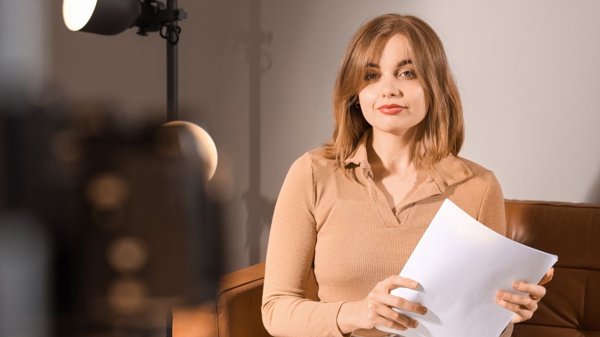 An actor sitting under a studio lamp, holding her sides and looking straight at the camera