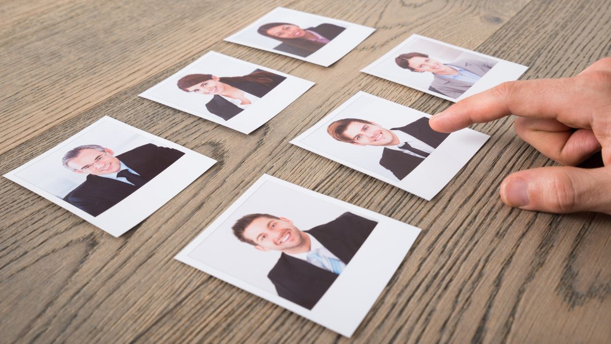 A hand reaching across six printed headshots laid out on a wooden table, moving one of them