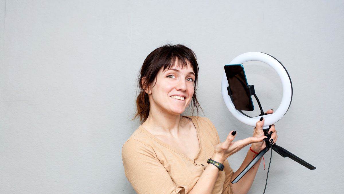 An actor filming herself on a phone clipped into a ring light, against a plain pale wall